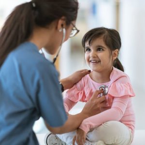 A toddler girl sits on a table during a medical examination. A female medical professional is using a stethoscope to listen to her heartbeat. The child is smiling up at her doctor.
